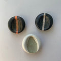 Three worry stones, one black with an orange, one black with a cream stripe, one white with a textured sage leaf impression in green.