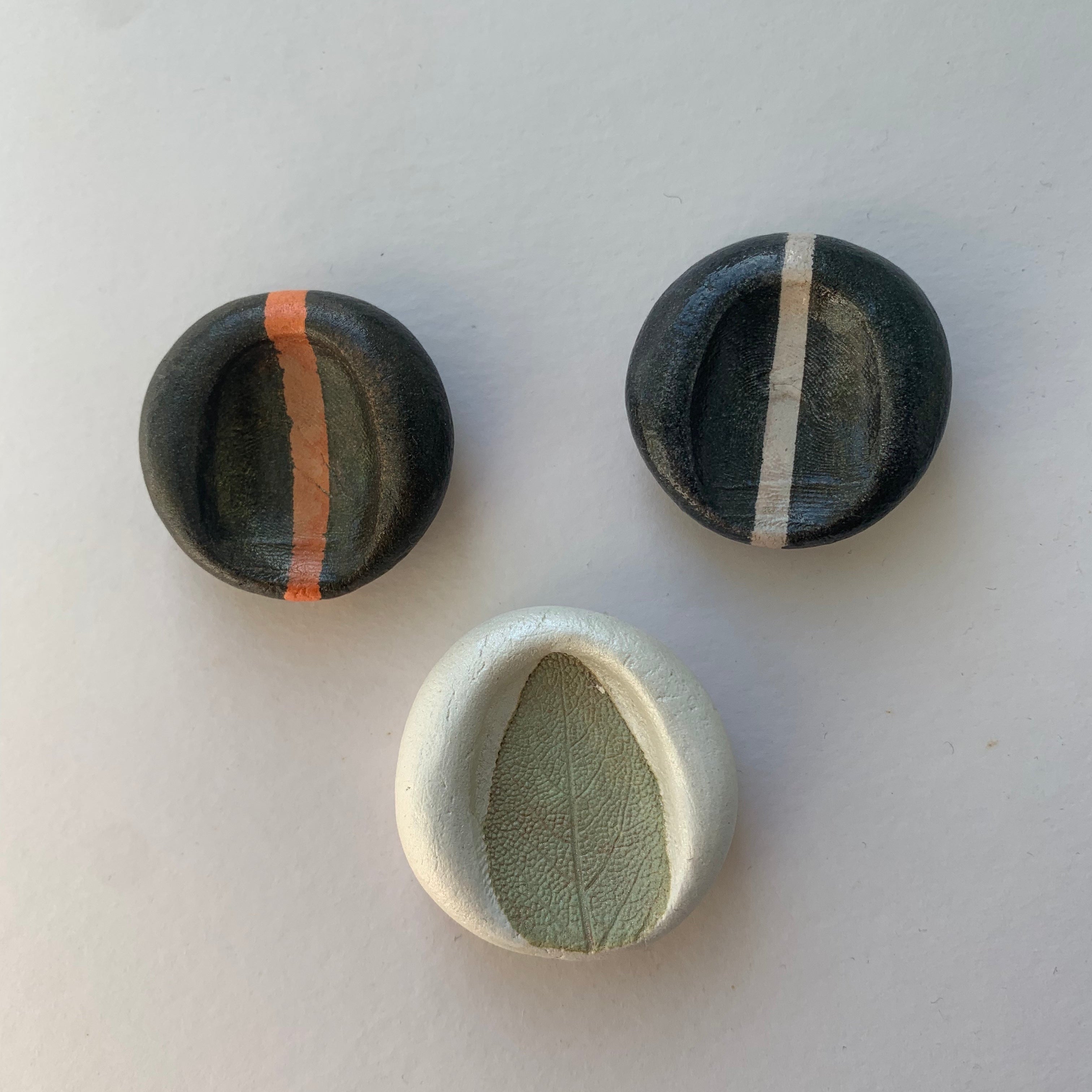 Three worry stones, one black with an orange, one black with a cream stripe, one white with a textured sage leaf impression in green.